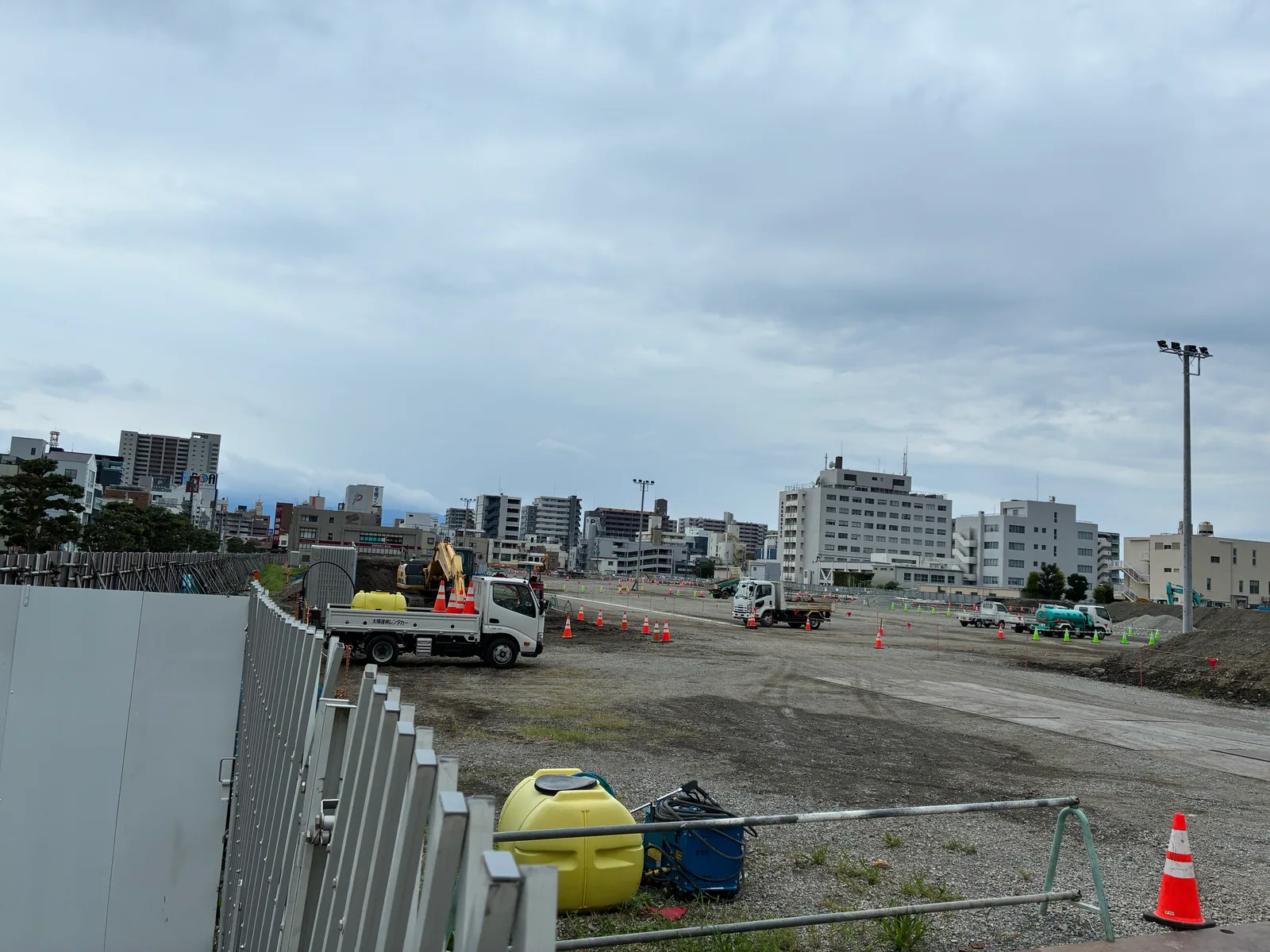 イオンタウン沼津の工事現場。広い更地に敷鉄板が敷かれ、カラーコーンと工事用トラック、散水車が並ぶ。奥に沼津駅北側のビル群