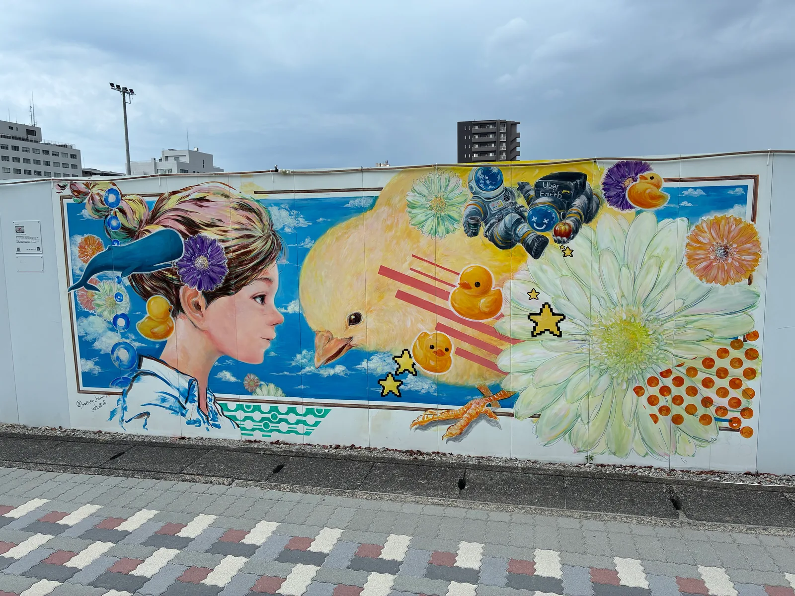 青空を背景に、花を髪に飾った子ども、大きなひよこ、アヒルのおもちゃ、宇宙飛行士、白い大輪の花が描かれた壁画「始まりについて」