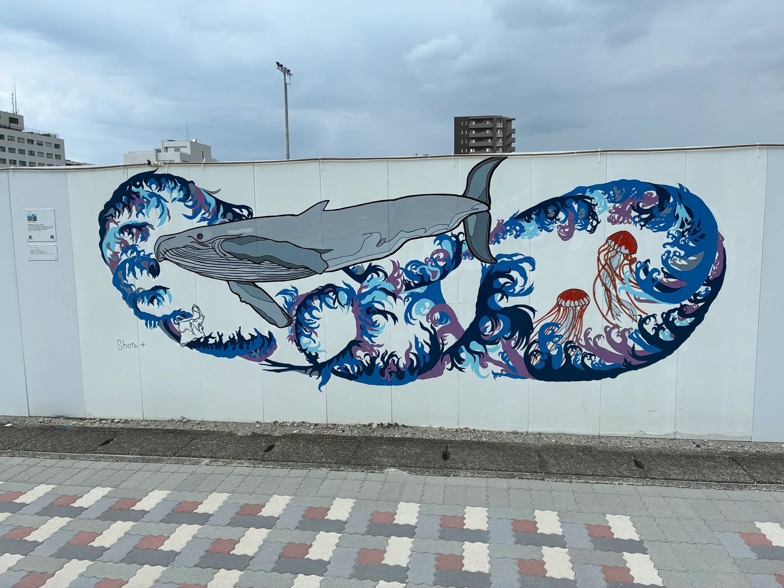 白い壁面に、青い波の渦の上を泳ぐザトウクジラと2匹のアカクラゲ、波間に漂うレジ袋が描かれた壁画「波とクジラとレジ袋」