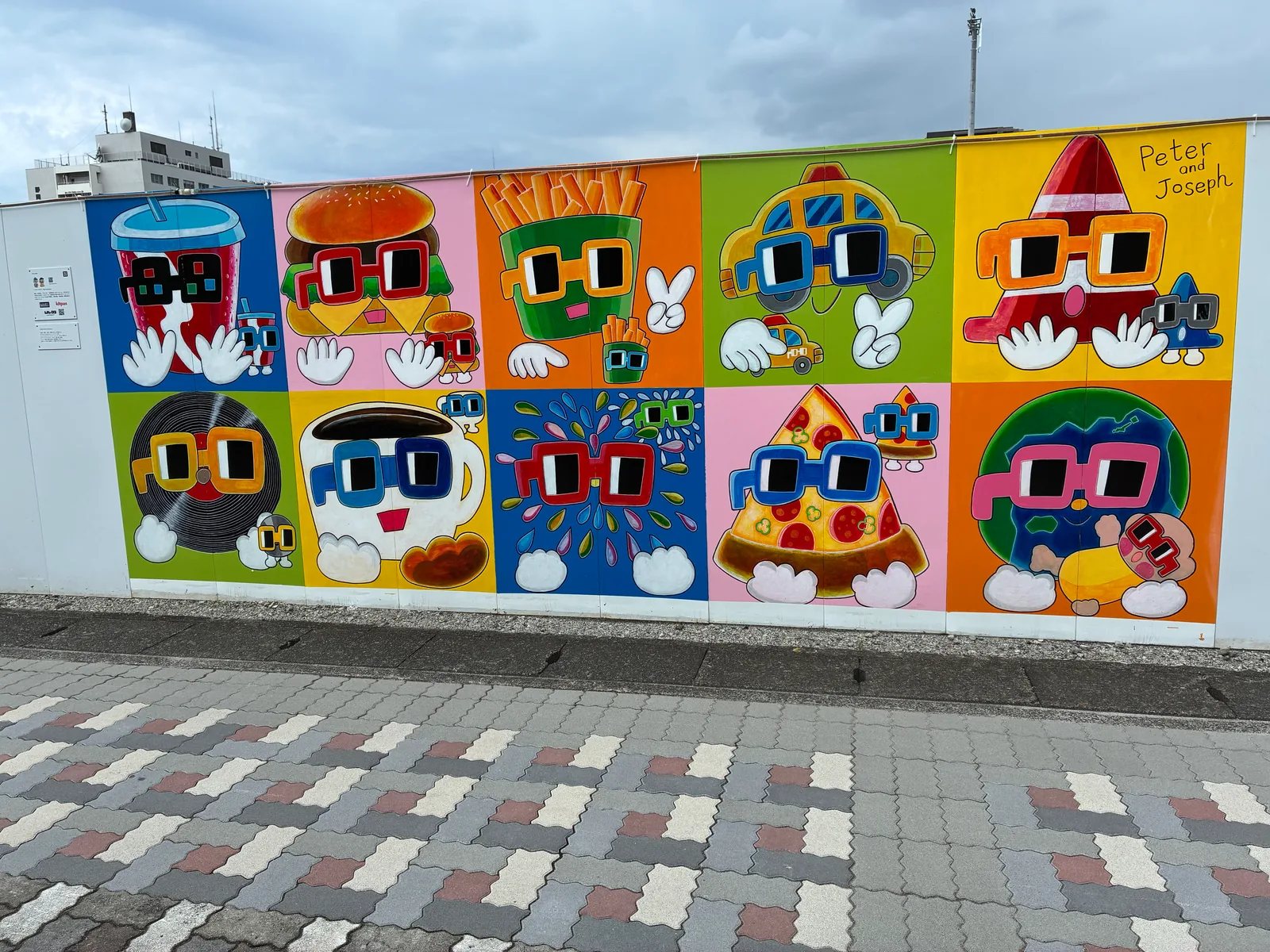 10色のマスに、大きなメガネをかけたドリンク、ハンバーガー、ポテト、タクシー、三角コーン、レコード、コーヒー、噴水、ピザ、地球のキャラクターが描かれた壁画