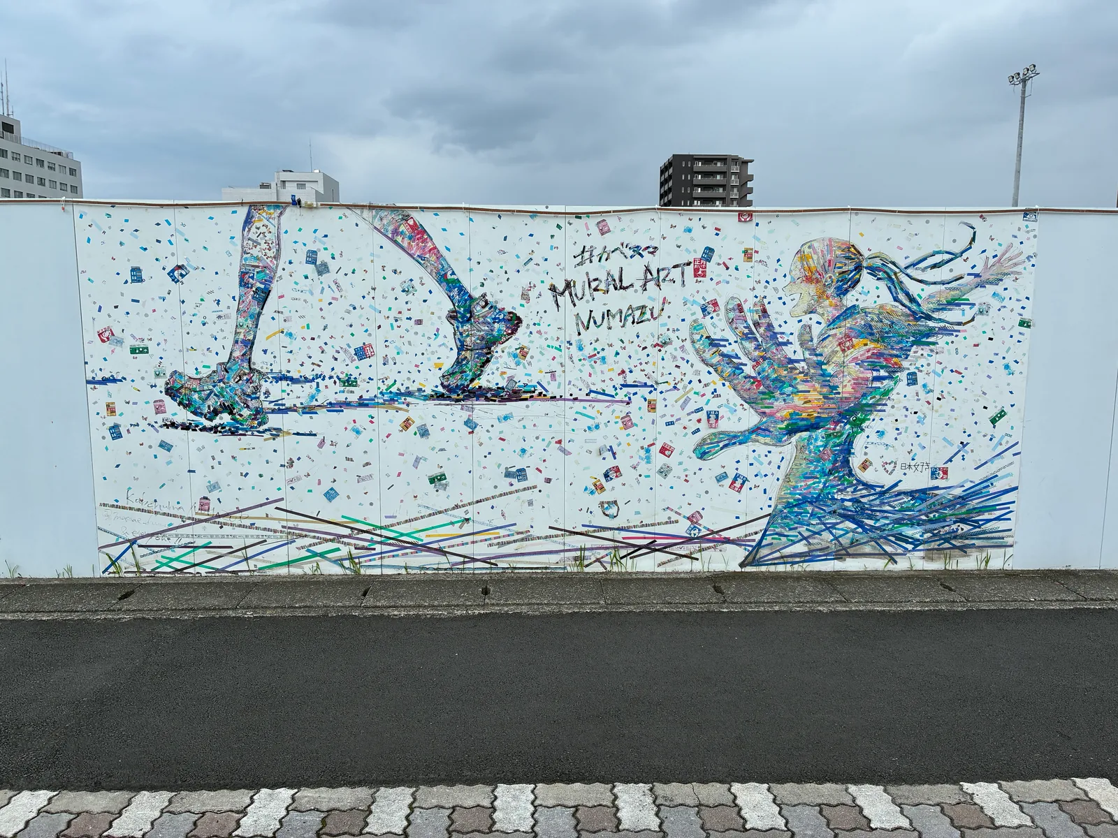 白い壁面に、色とりどりのマスキングテープやシールを貼り重ねて描いた走る脚と、手を伸ばして駆ける少女。中央に「#カベヌマ MURAL ART NUMAZU」の文字
