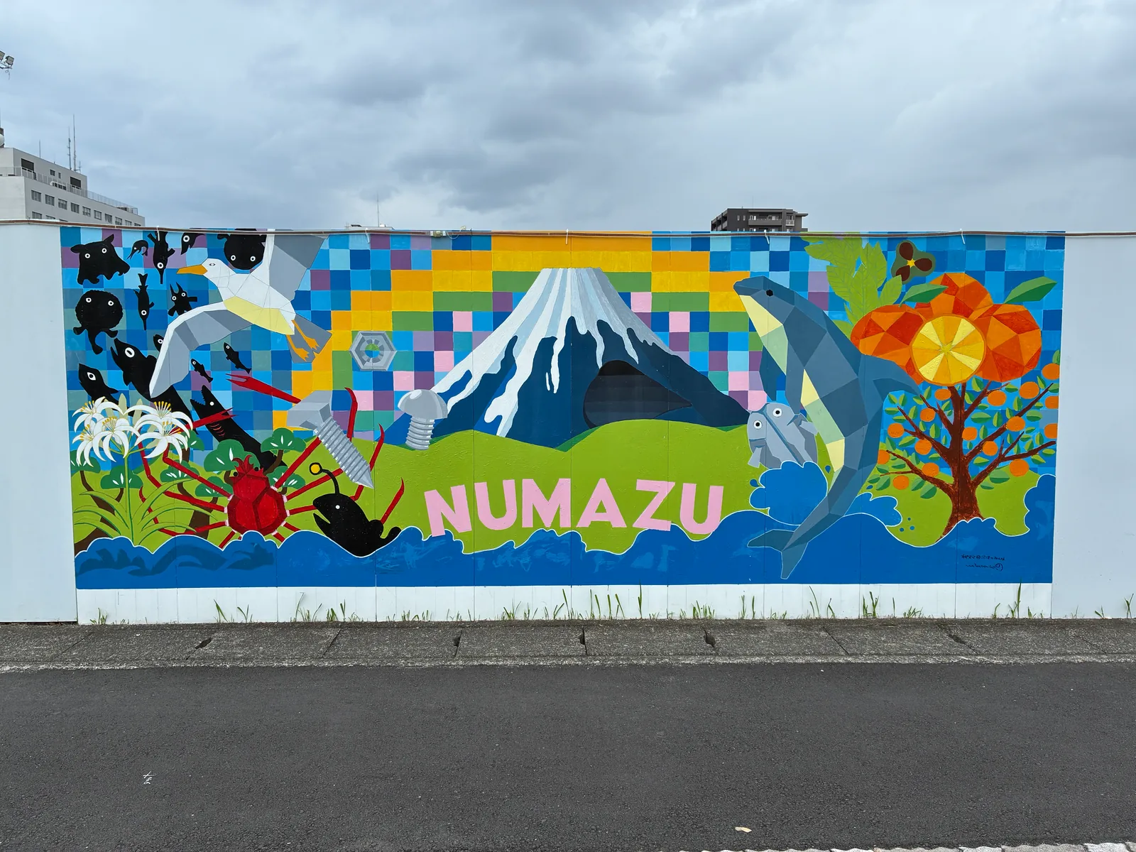 カラフルなモザイク柄の空を背景に、富士山、ピンクの「NUMAZU」の文字、深海魚のシルエット、タカアシガニ、カモメ、イルカ、みかんの木、ハマユウが描かれた壁画
