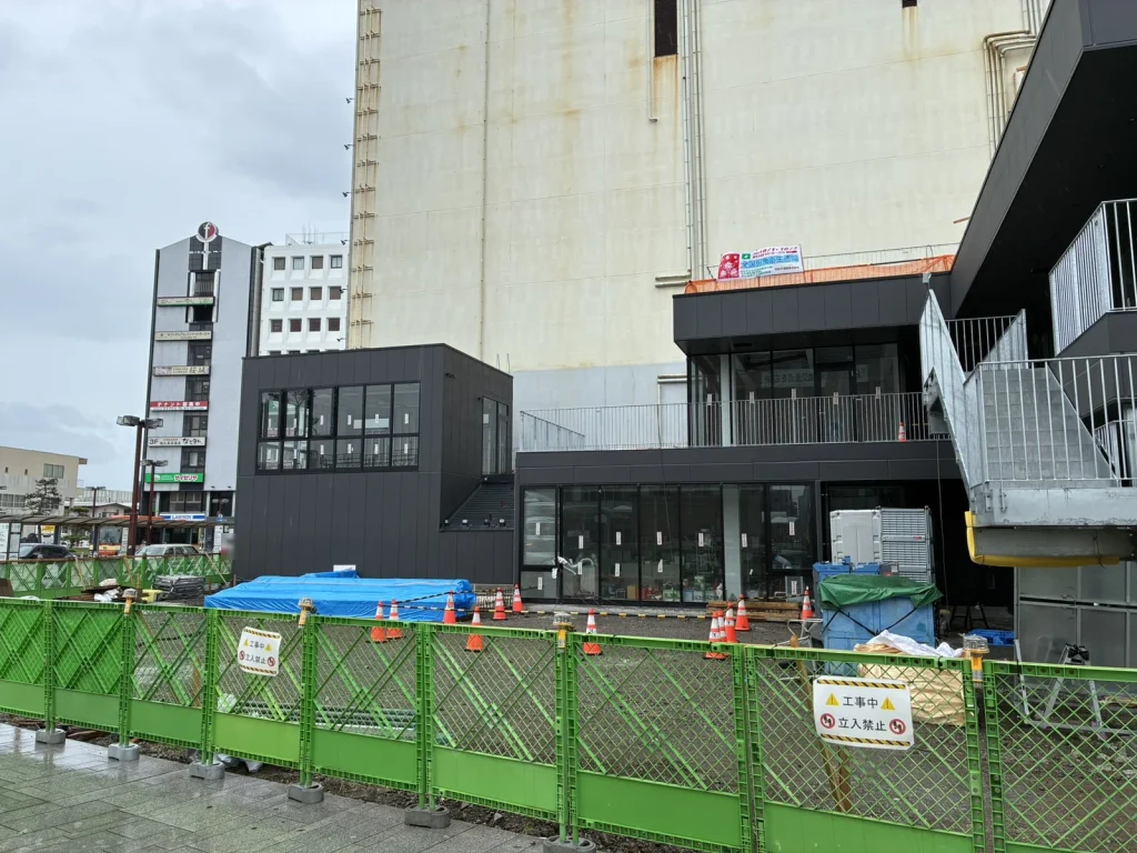 NUMAZU JAMSの建物脇、まだ土のままの広場予定地。緑の仮囲いとカラーコーンが並ぶ(2026年9月・沼津駅南口)
