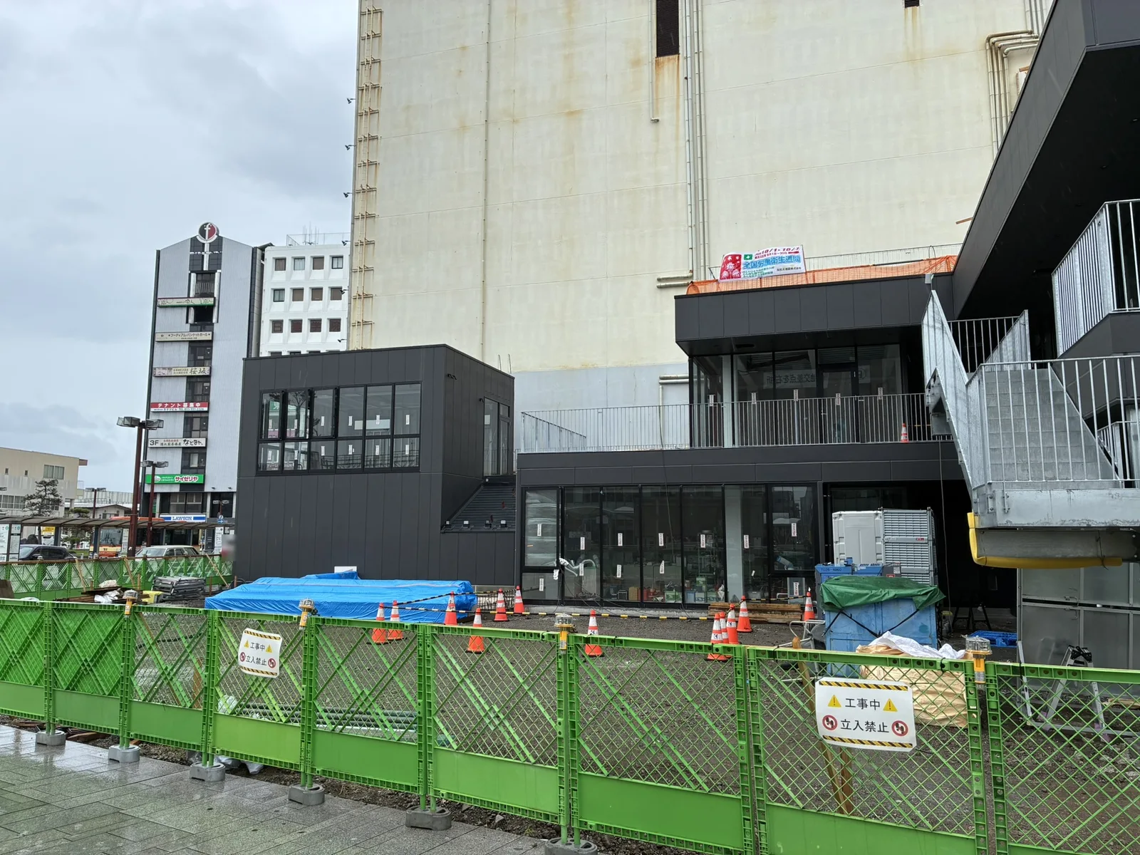 NUMAZU JAMSの建物脇、まだ土のままの広場予定地。緑の仮囲いとカラーコーンが並ぶ(2026年9月・沼津駅南口)