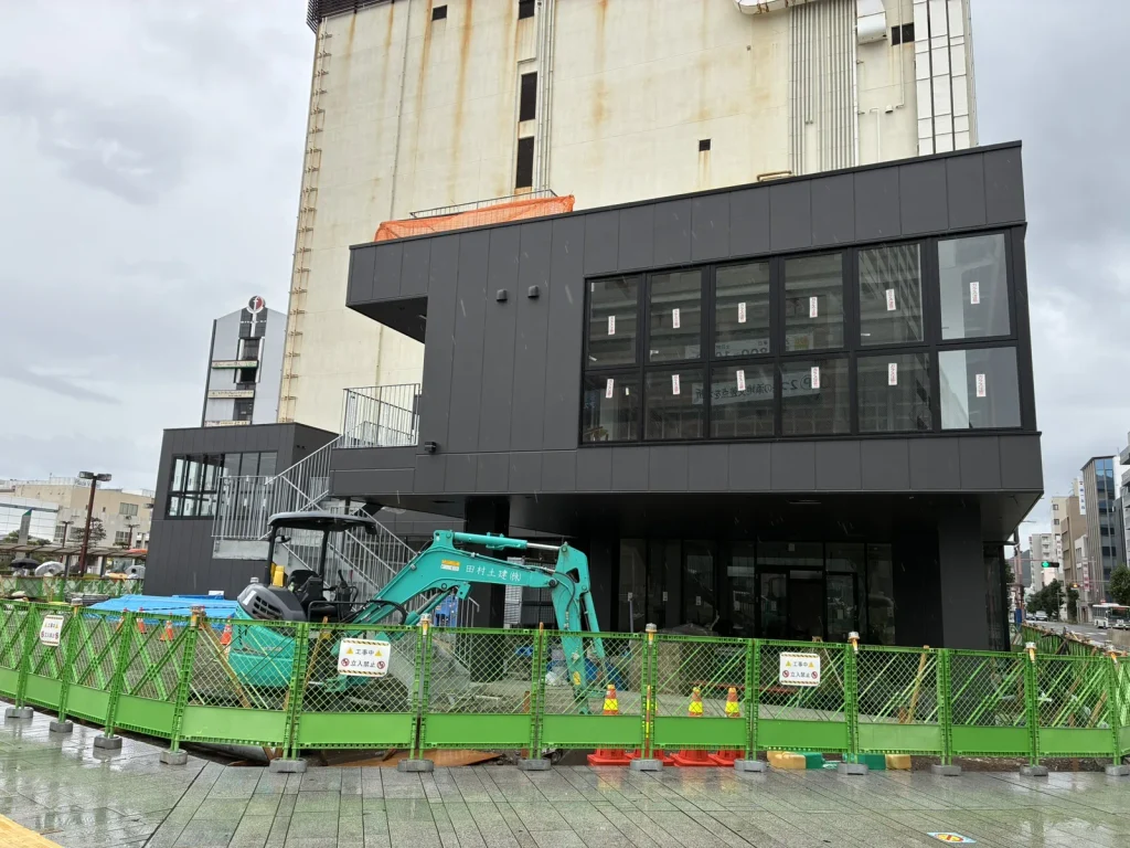 別角度から見たNUMAZU JAMSの建物と、足元で作業する小型重機。背後は旧西武百貨店沼津店の建物(2026年9月・沼津駅南口)