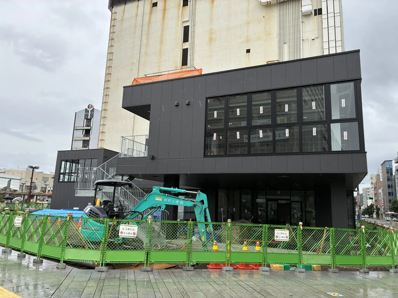別角度から見たNUMAZU JAMSの建物と、足元で作業する小型重機。背後は旧西武百貨店沼津店の建物(2026年9月・沼津駅南口)