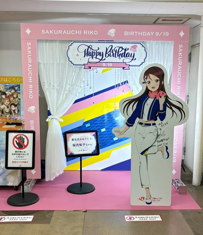 「SAKURAUCHI RIKO BIRTHDAY 9/19」と書かれたピンクの枠。中にレースのカーテンと「Happy Birthday 9.19」の看板、手前に桜内梨子の等身大パネルと「誕生日おめでとう！桜内梨子ちゃん JR東海より」の立て看板