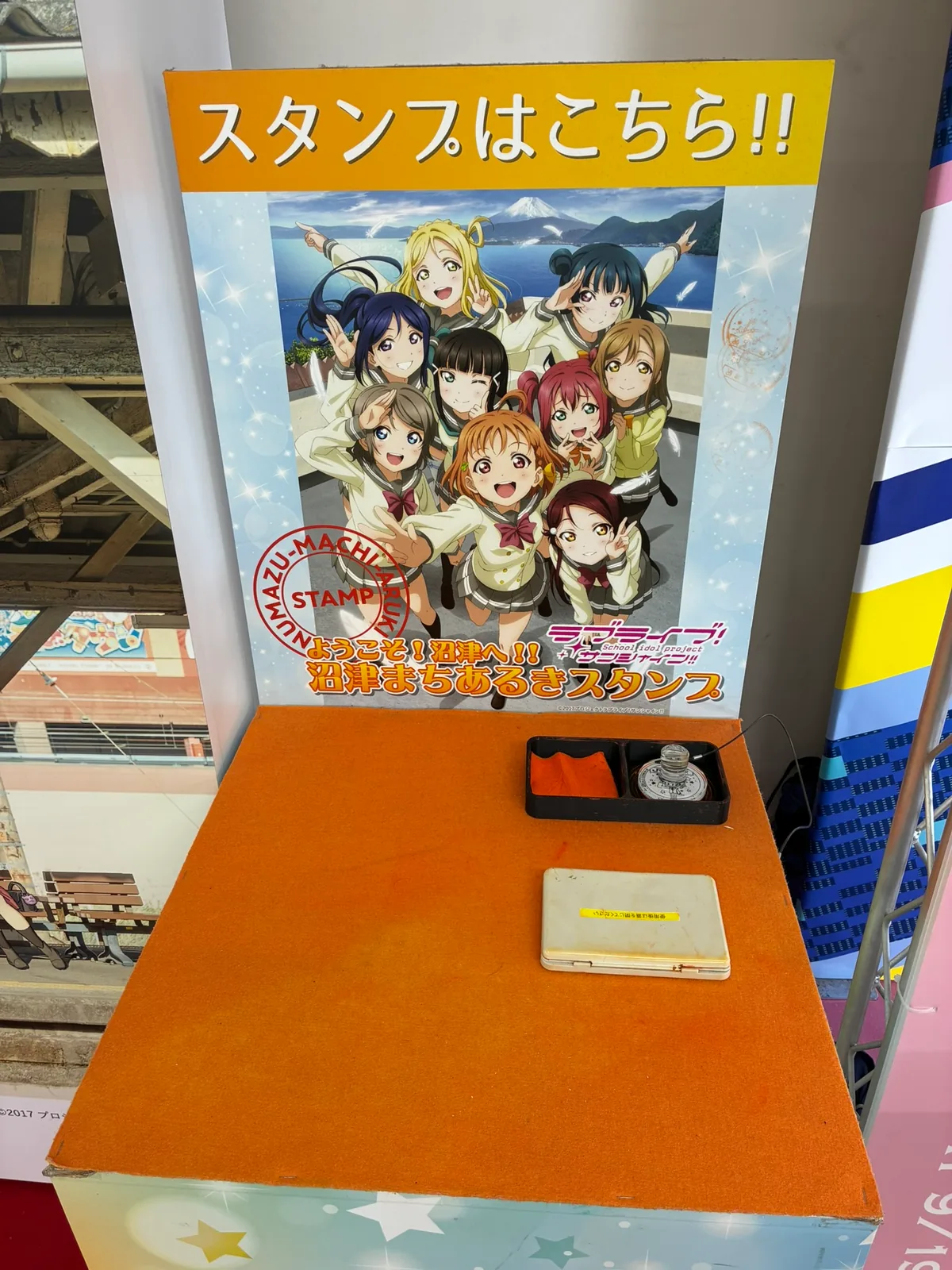 「スタンプはこちら!!」の看板とAqours9人のイラスト。オレンジのフェルト張りの台にスタンプとスタンプ台が置いてある