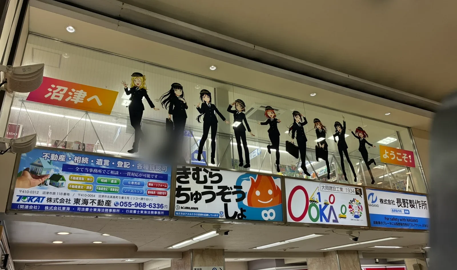 沼津駅南口のコンコース。2階のガラス面に駅員の制服姿のAqours9人のパネルが並び、左に「沼津へ」、右に「ようこそ！」。下に地元企業の広告看板