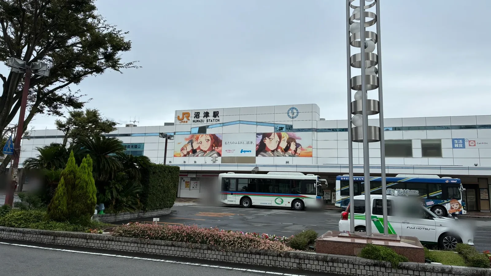 ロータリーの向かいから見た沼津駅南口。駅舎に「私たちのふるさと、沼津。」とAqoursの幕、手前に伊豆箱根バスとラブライブ！サンシャイン!!のラッピングバス、タクシー