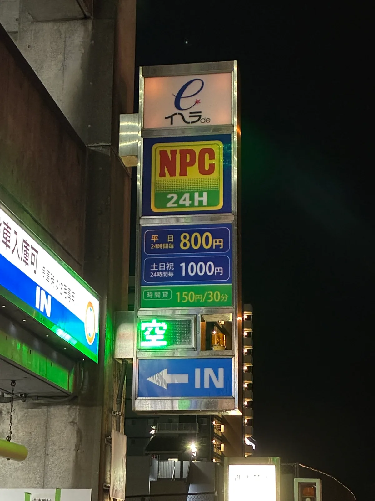 NPC24Hイーラdeパーキングの入口の袖看板。平日24時間毎800円、土日祝24時間毎1000円、時間貸150円/30分、満空表示は「空」、左向きの矢印とIN