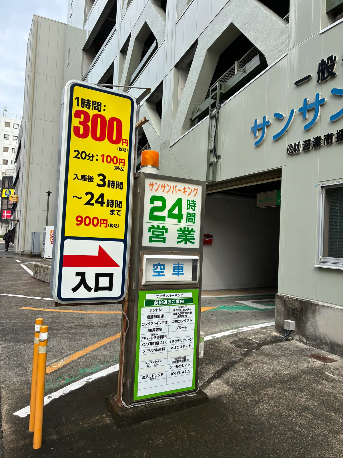 サンサンパーキングの入口。1時間300円、20分100円、入庫後3時間から24時間まで900円、24時間営業、空車表示、契約店の一覧。奥に立体駐車場の建物