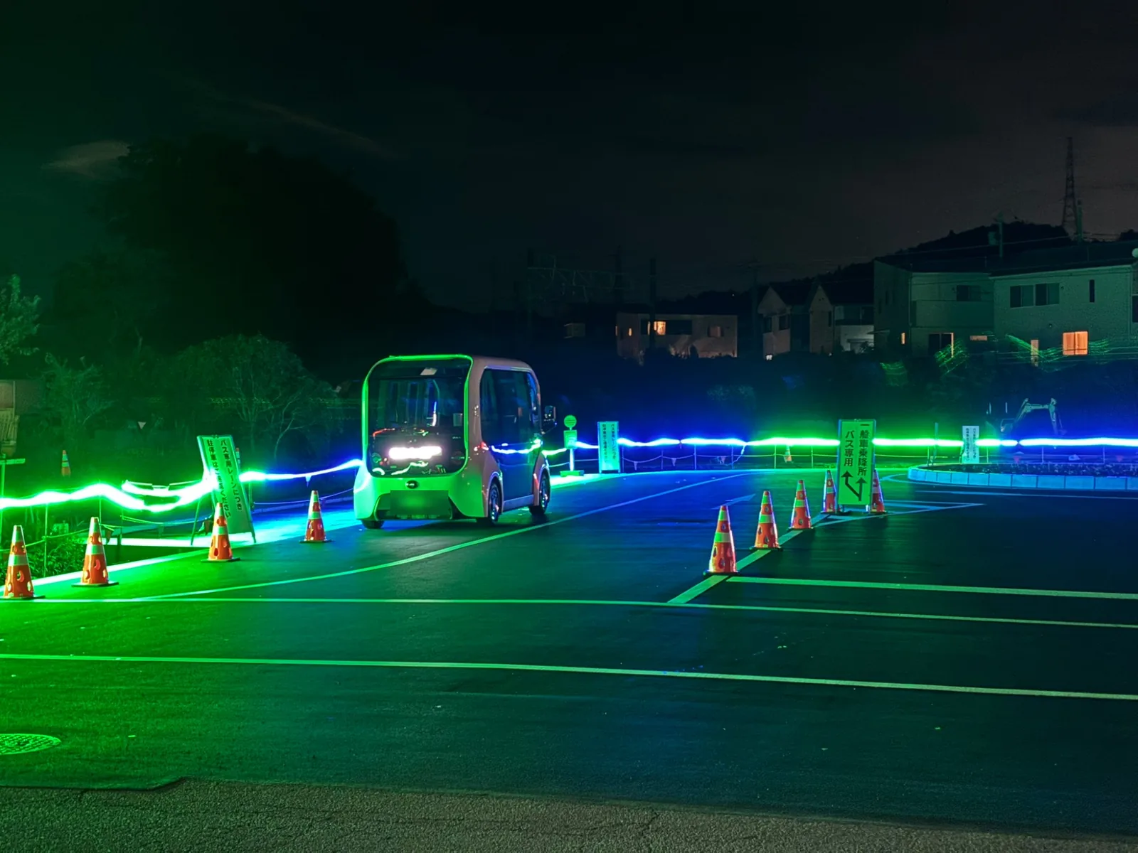 夜の岩波駅前ロータリー。バス専用レーンに入ってくる箱形の小型EVバス。仮囲いに沿って青と緑のラインライトが灯っている