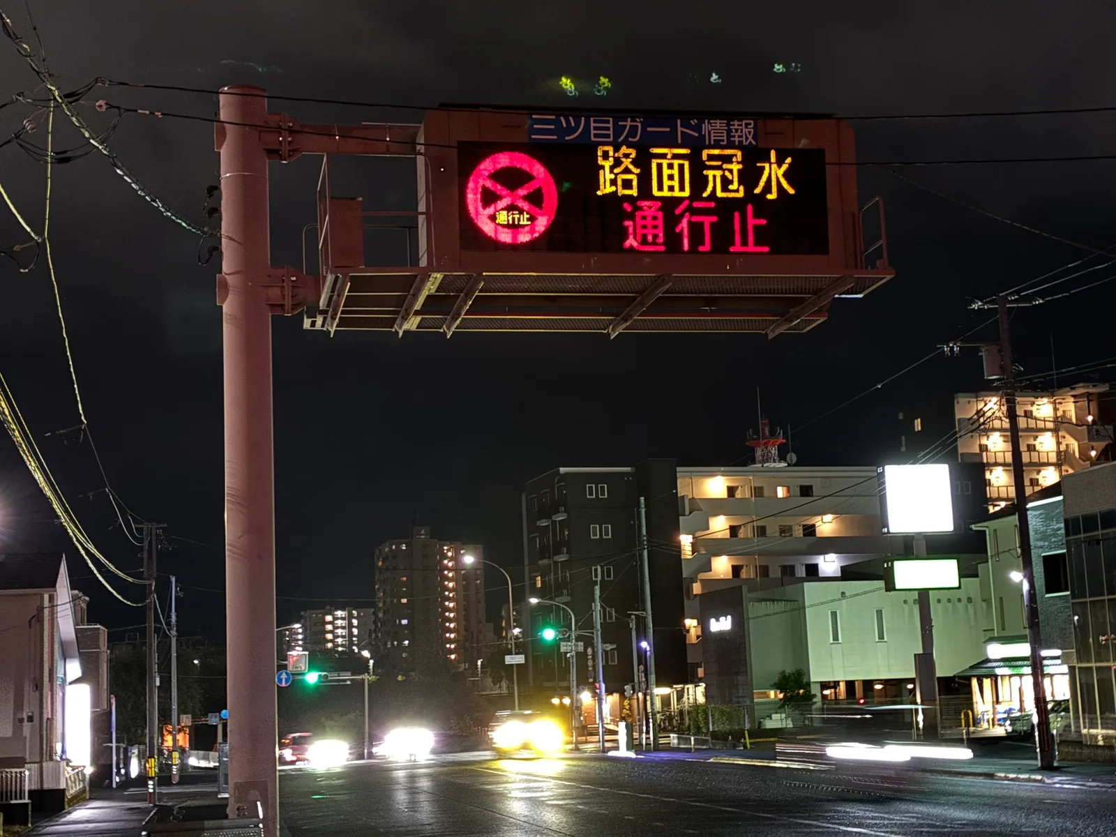 夜の交差点の上に掛かる「三ツ目ガード情報」の電光掲示板。赤い通行止めのマークと「路面冠水 通行止」の文字が光り、下の道路は雨で濡れている