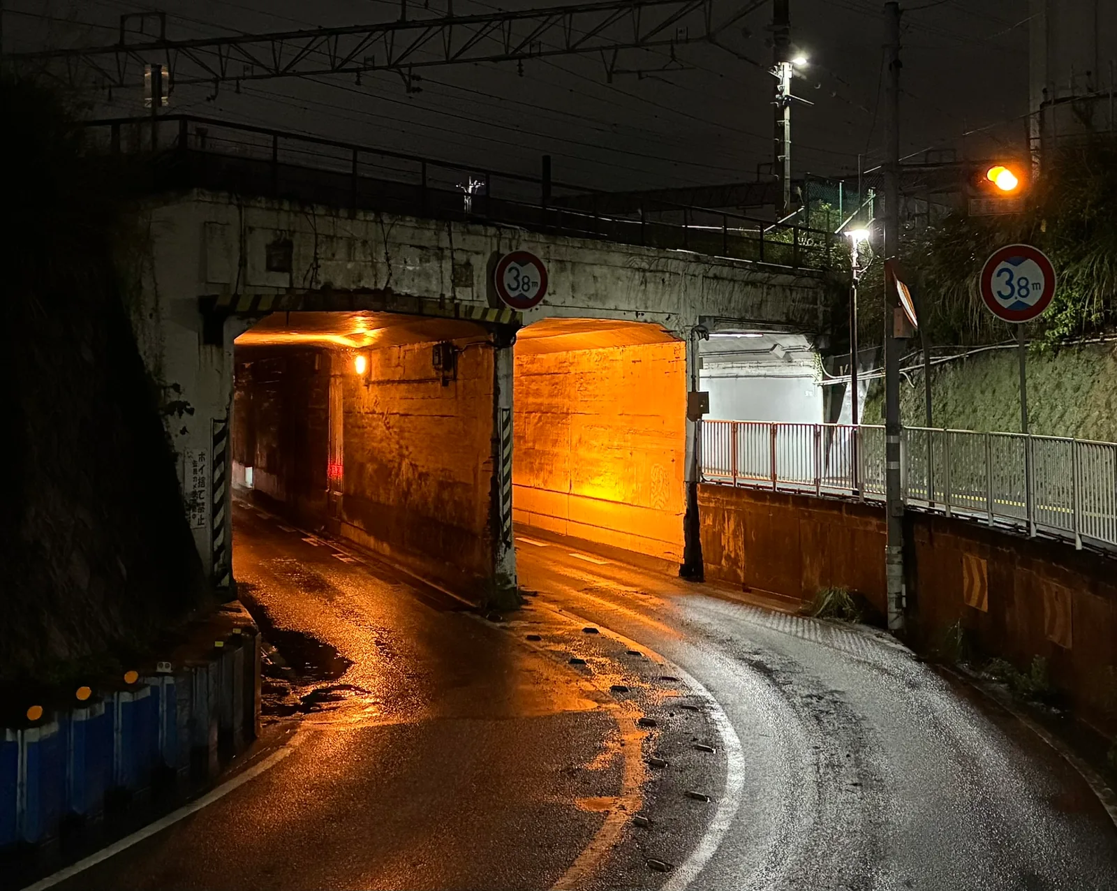 線路の下をくぐるコンクリートのガード。2つに分かれた開口部がオレンジ色の照明で照らされ、高さ3.8mの制限標識が掛かっている。手前の路面には水たまりが残る
