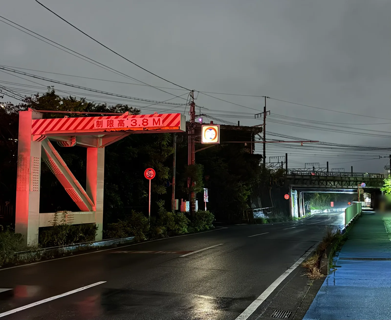 曇った夕方の国道414号。左に赤白の縞模様の門型「制限高3.8M」が立ち、まっすぐ延びる道路の先に線路の高架橋とガードの入口が見える