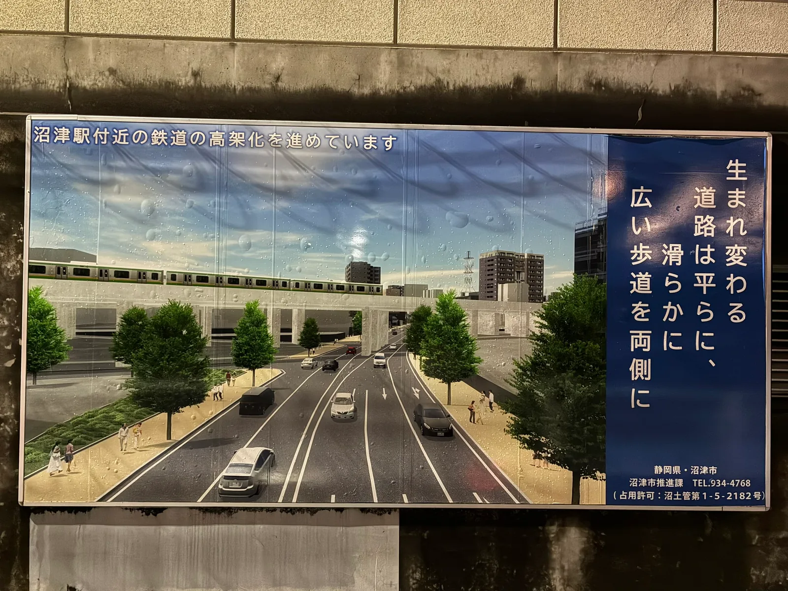 ガード下の壁に貼られた掲示。高架を走る電車の下に、並木と広い歩道のある平らな4車線道路が描かれ、「生まれ変わる道路は平らに、滑らかに、広い歩道を両側に」と書かれている