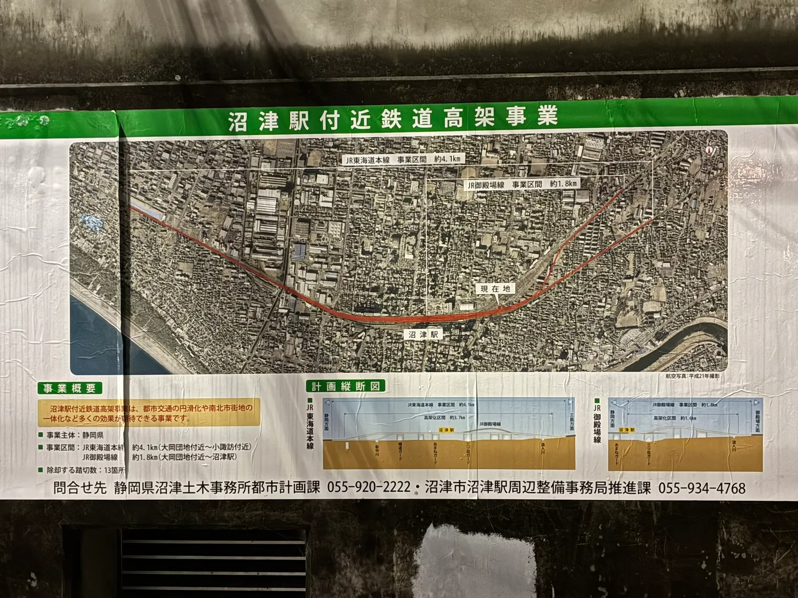 「沼津駅付近鉄道高架事業」の掲示。航空写真に高架化する東海道本線と御殿場線が赤い線で描かれ、下に事業概要と、三ツ目ガードの位置が入った計画縦断図がある